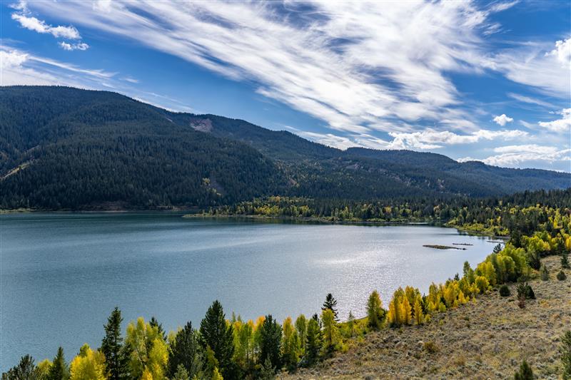 Tranquil mountain lake bordered by forested hills and distant mountains under a partly cloudy sky