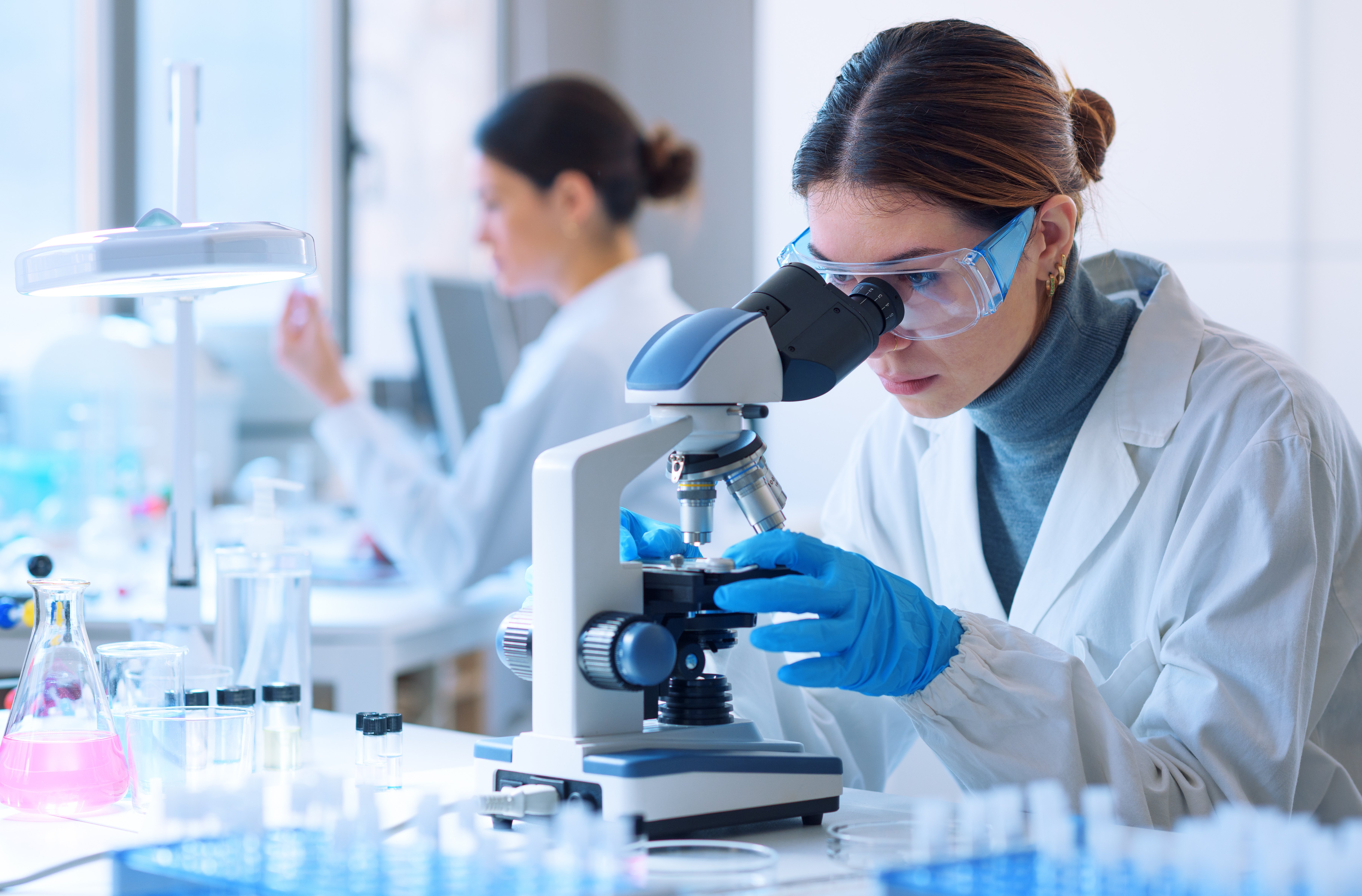A female scientist conducts research in a laboratory setting while a coworker works in the background.