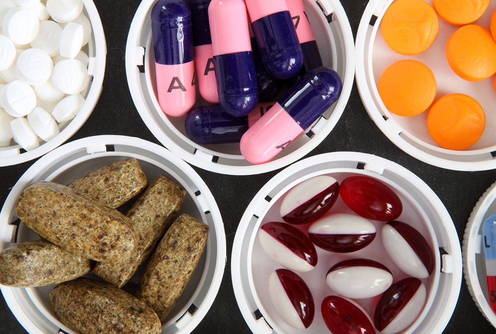 The image shows a variety of pills in different shapes, sizes, and colors organized in plastic containers.
