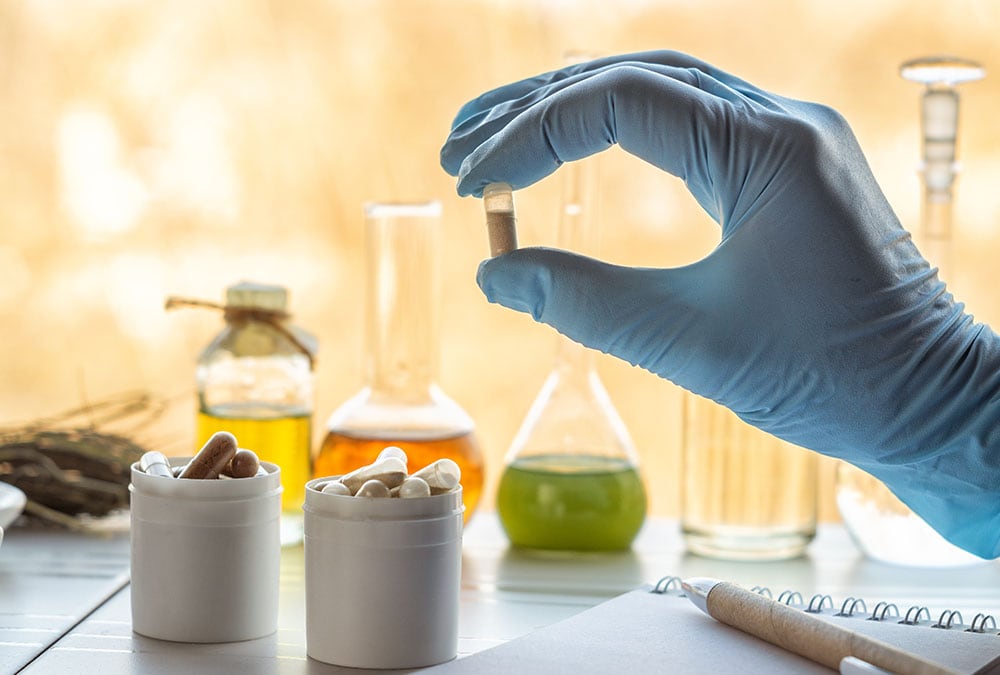 A hand in a blue glove holds a capsule while other capsules and lab equipment are in the background.