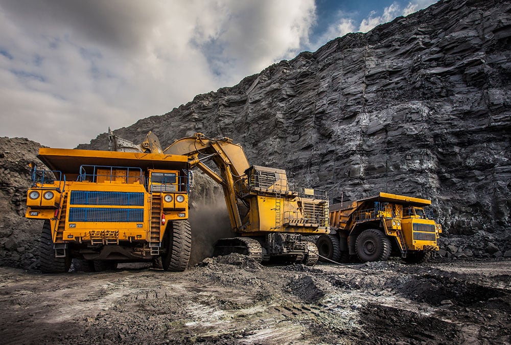 Two large mining trucks and a mining excavator are seen against a backdrop of a coal quarry.