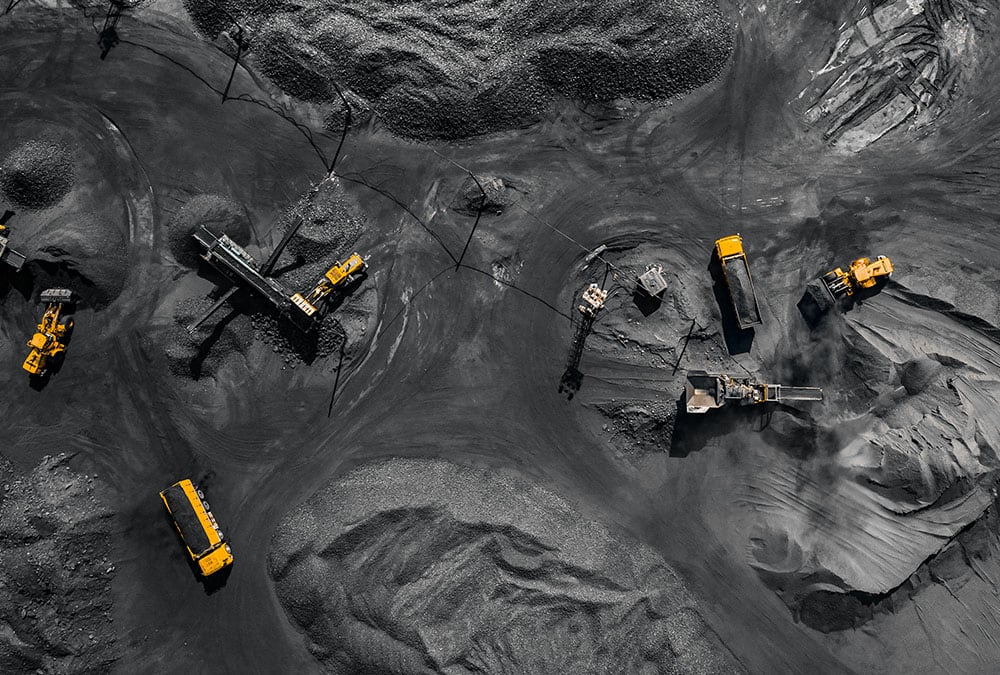 Several pieces of yellow heavy machinery are working on a large, flat, grey area in an aerial view.