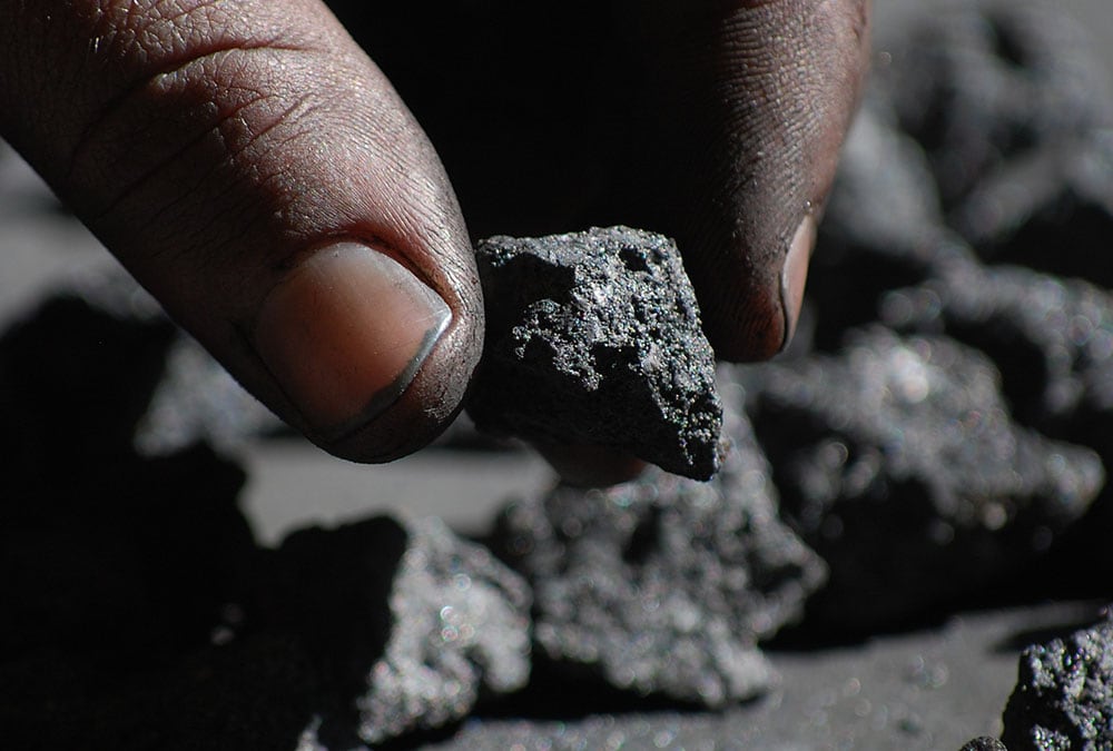 A hand holds a piece of coal, with more coal in the background.
