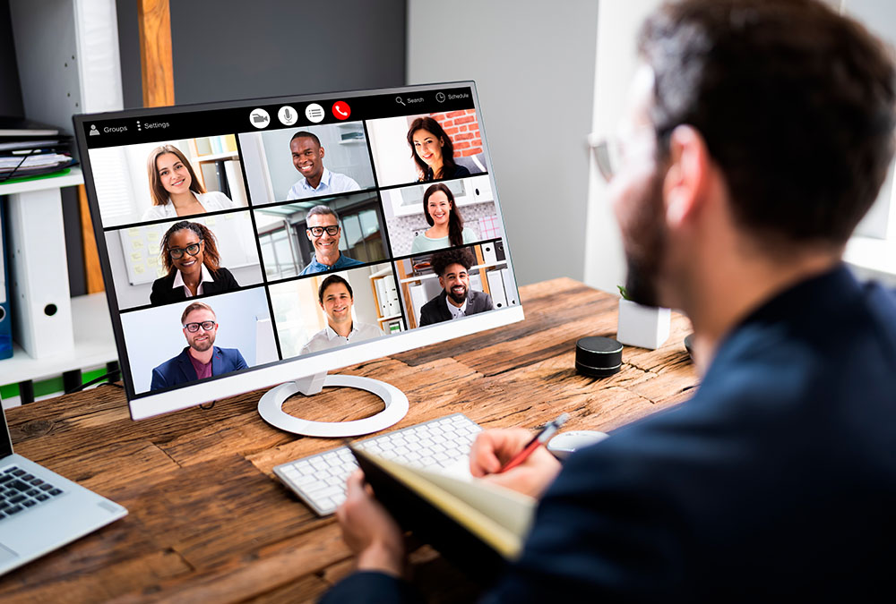 A man is on a video conference call with his team while writing notes in a notebook.