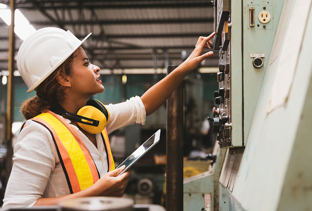 A woman in a hard hat and safety vest adjusts a machine control panel while holding a tablet.