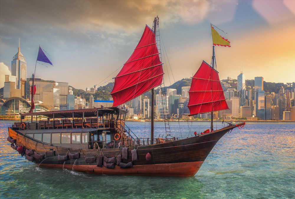 A traditional Chinese junk ship with red sails anchors in a bay in front of a busy city skyline.