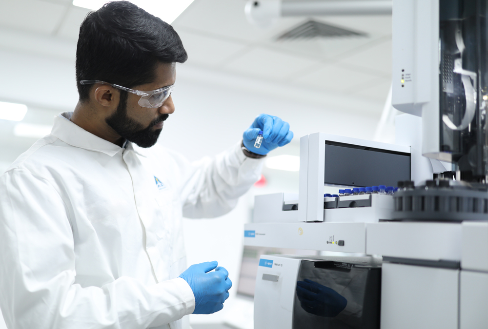 A lab technician wearing a lab coat and protective glasses inserts a vial into a testing device.