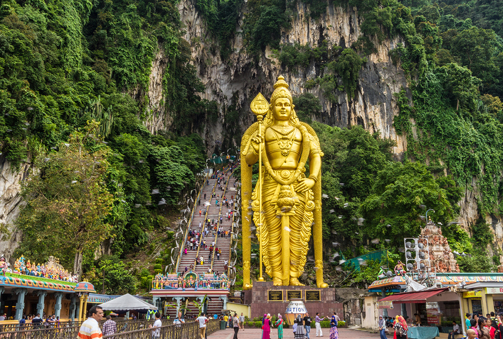 The image shows a large golden statue of a Hindu deity, standing in front of a cave with steps leading up to an entrance.