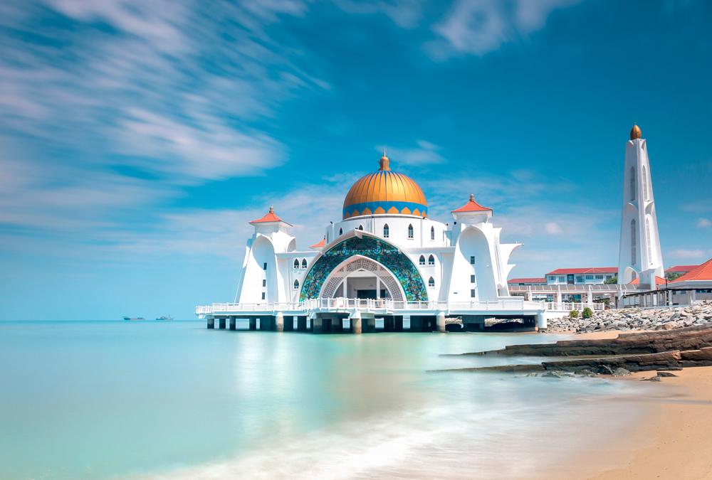 A white mosque with a golden dome sits on a pier above the calm, turquoise water.