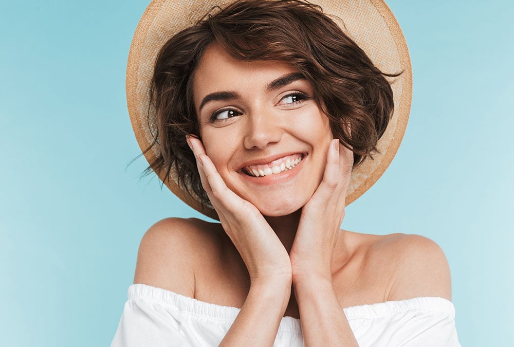 A young smiling woman with short brown hair is wearing a straw hat and a white off-the-shoulder top.