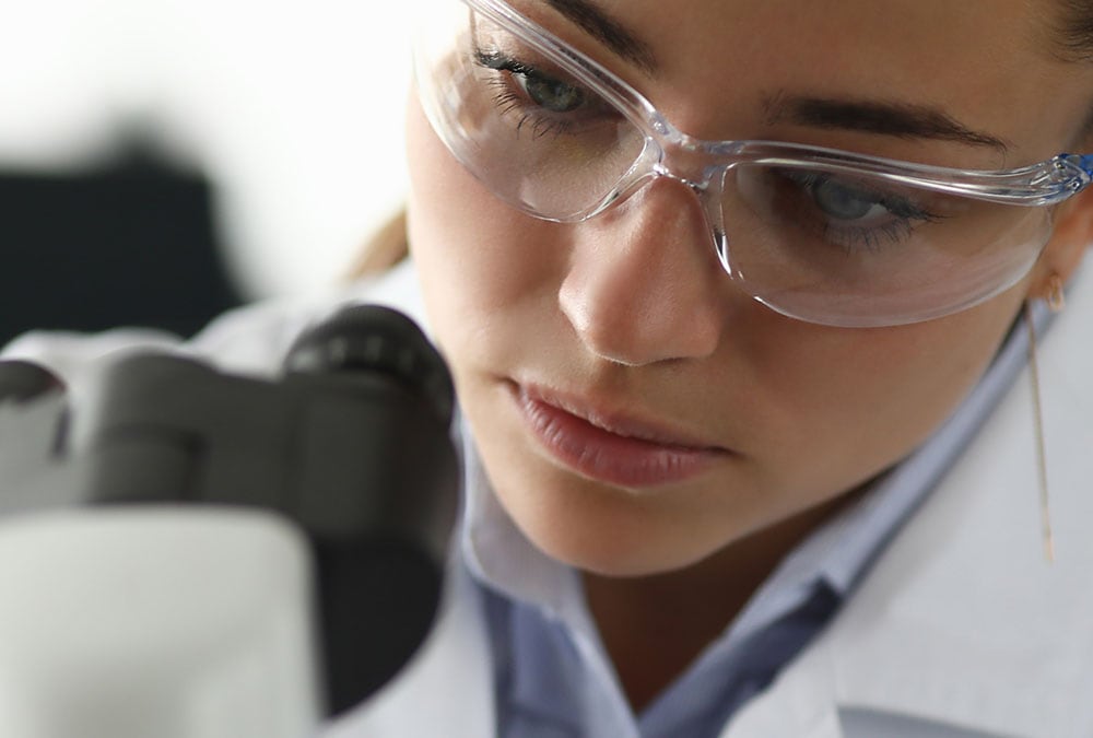 A woman in a lab coat and safety goggles is looking intently through a microscope.