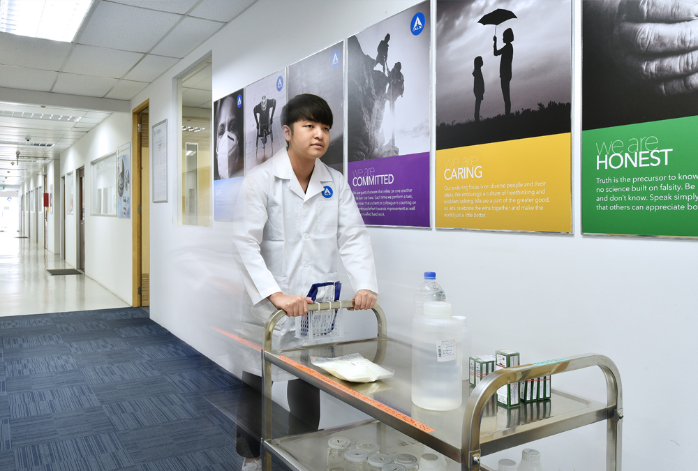 A lab technician pushes a cart with laboratory equipment down a hallway with posters about company values lining the walls.