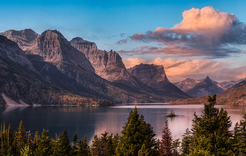 A lake surrounded by mountains and evergreen trees, with a cloud filled evening sky.