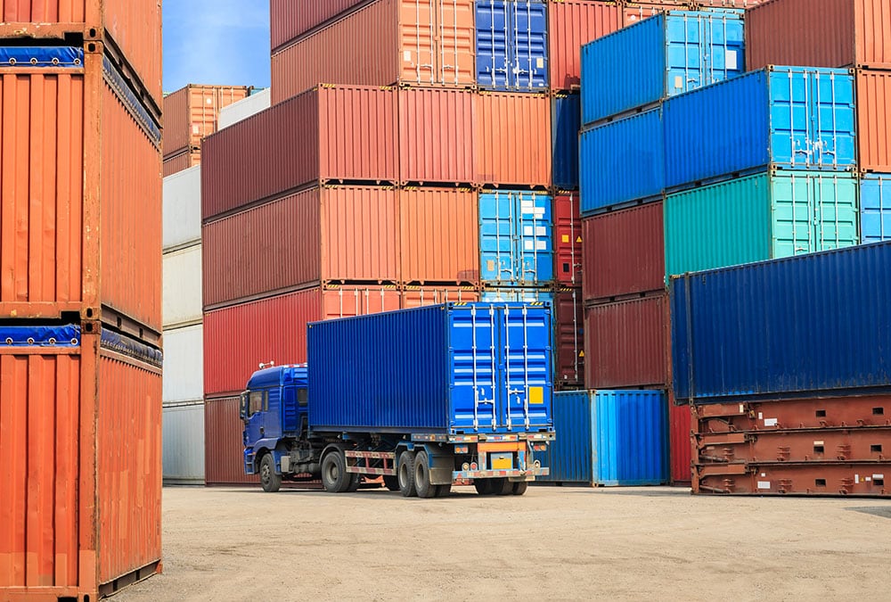 A blue delivery truck is parked in front of a wall of stacked shipping containers.