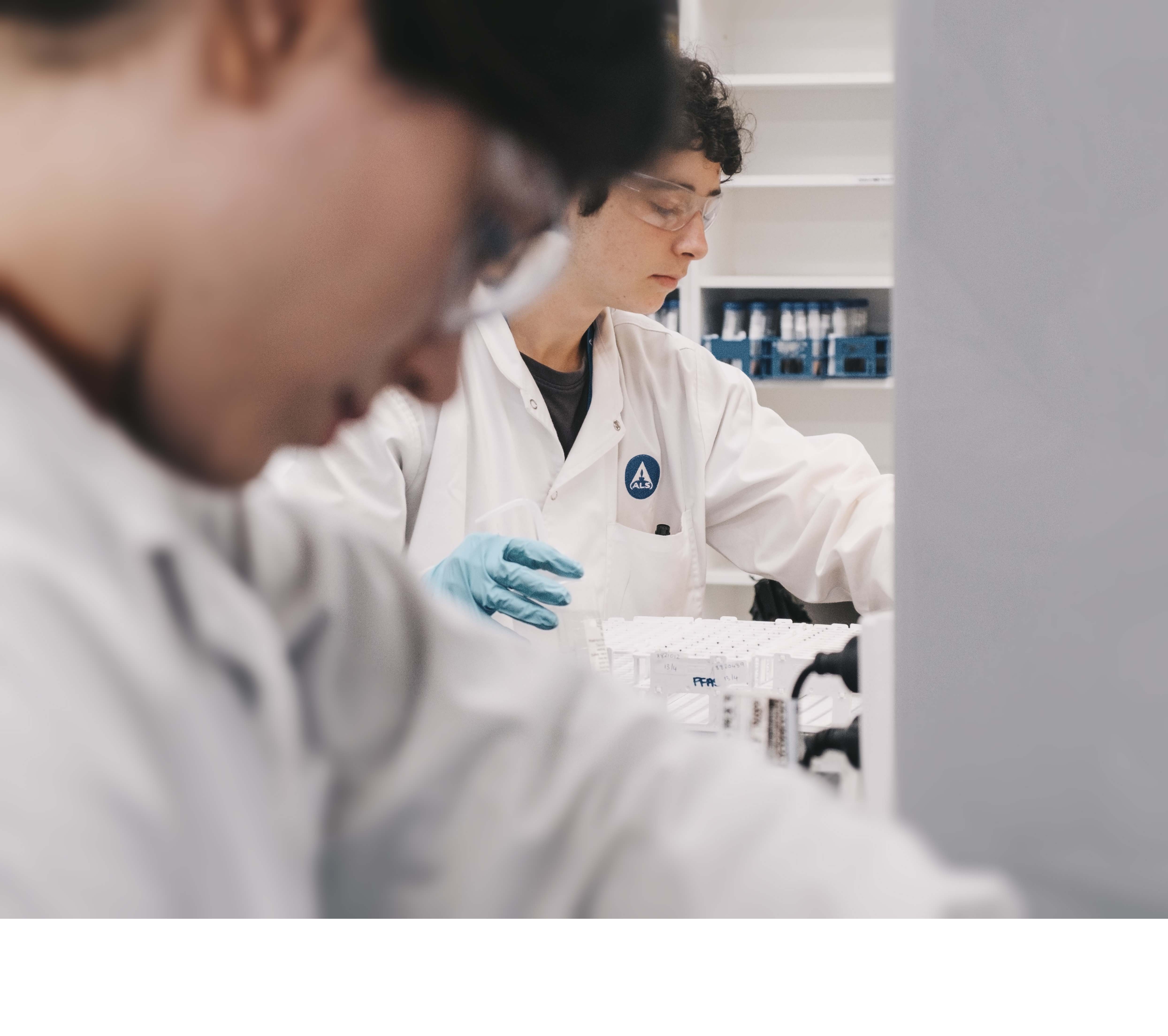 Researchers in a laboratory wearing white lab coats and safety glasses, handling scientific equipment.