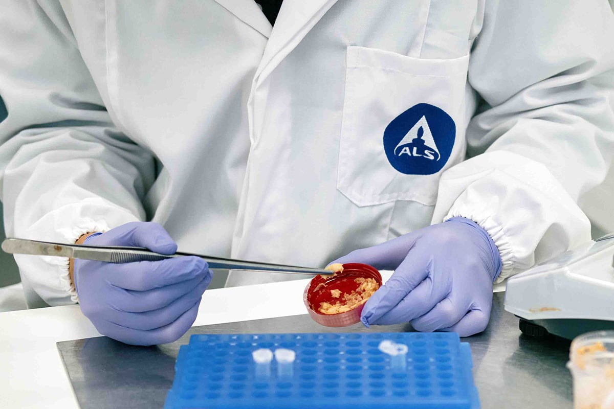 Person wearing a white lab coat with the "ALS" logo and purple gloves, using tweezers to handle a petri dish with a red substance. A blue rack with small test tubes is visible on the table.
