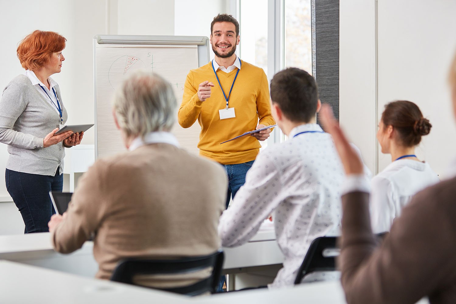 Sessão de Consultoria e Formação Profissional Grupo de pessoas a assistir a uma apresentação numa sala de formação. Uma pessoa está de pé à frente com um caderno, enquanto outra segura um tablet. O público está sentado e atento.