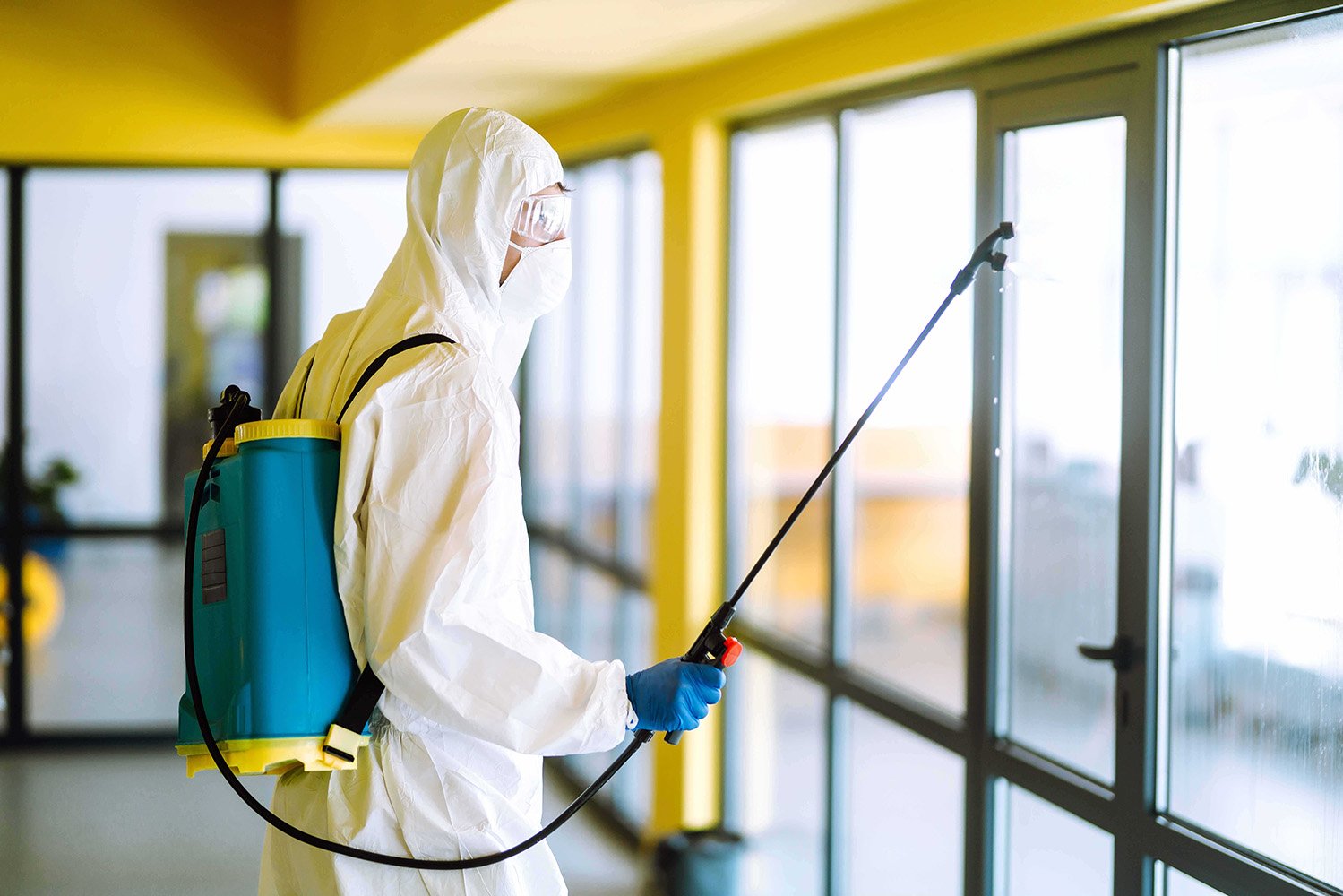Desinfeção Profissional com Equipamento de Proteção Individual Pessoa com fato de proteção completo, luvas e óculos, utilizando um pulverizador de mochila azul e amarelo para desinfetar portas e janelas de vidro num espaço interior com paredes amarelas.