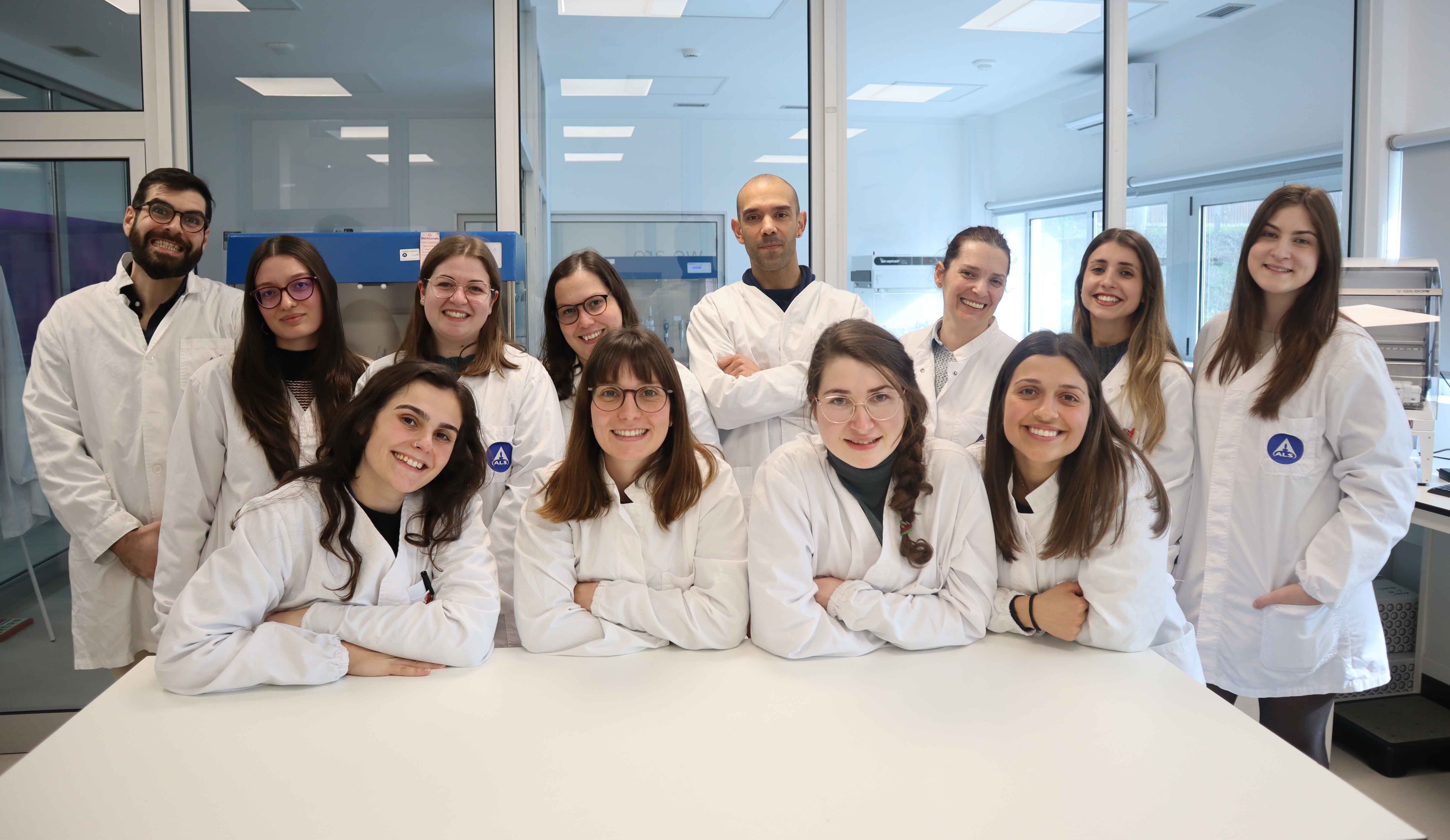 Grupo de Cientistas em Laboratório Grupo de pessoas com batas brancas sentadas e em pé à volta de uma mesa num ambiente de laboratório.