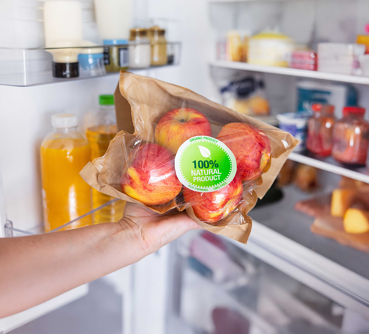 Mão a segurar um saco com maçãs embaladas rotulado como “100% produto natural”, em frente a um frigorífico aberto com vários alimentos no interior.