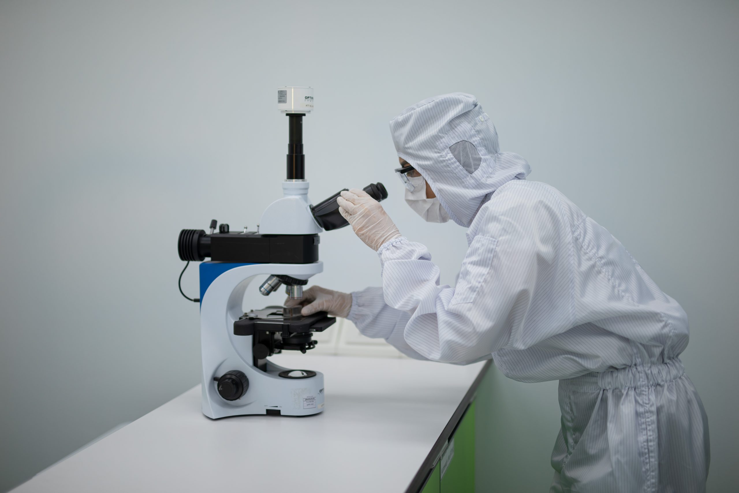 A person wearing cleanroom protective clothing uses a laboratory microscope to examine a sample on a workbench in a controlled laboratory environment.