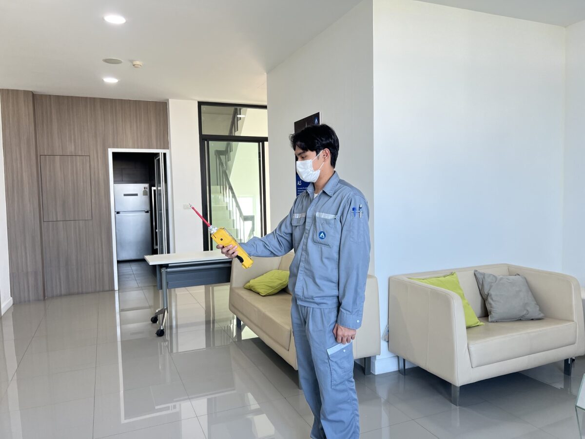 A person wearing a mask and work uniform uses a handheld air quality measuring device inside a modern indoor office or laboratory lounge area.