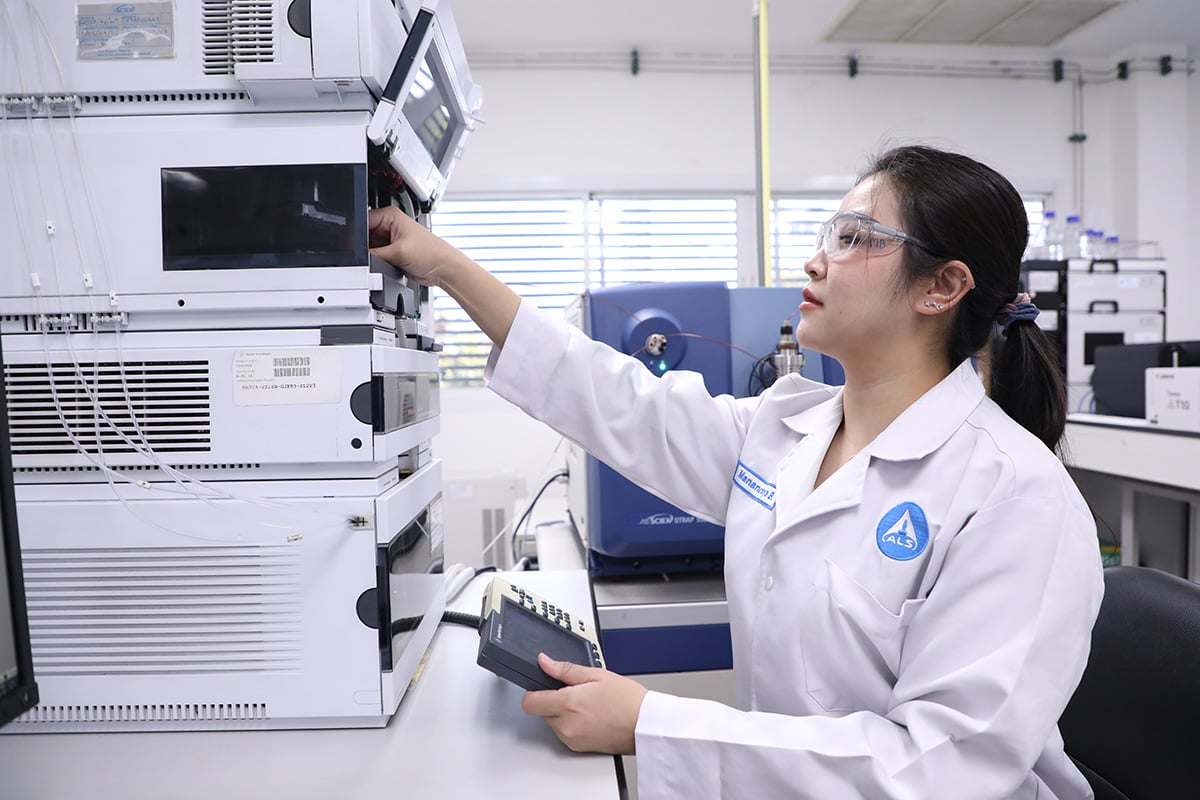 A person wearing a laboratory coat adjusts controls on a large stack of analytical laboratory equipment while holding a handheld device inside a laboratory environment.