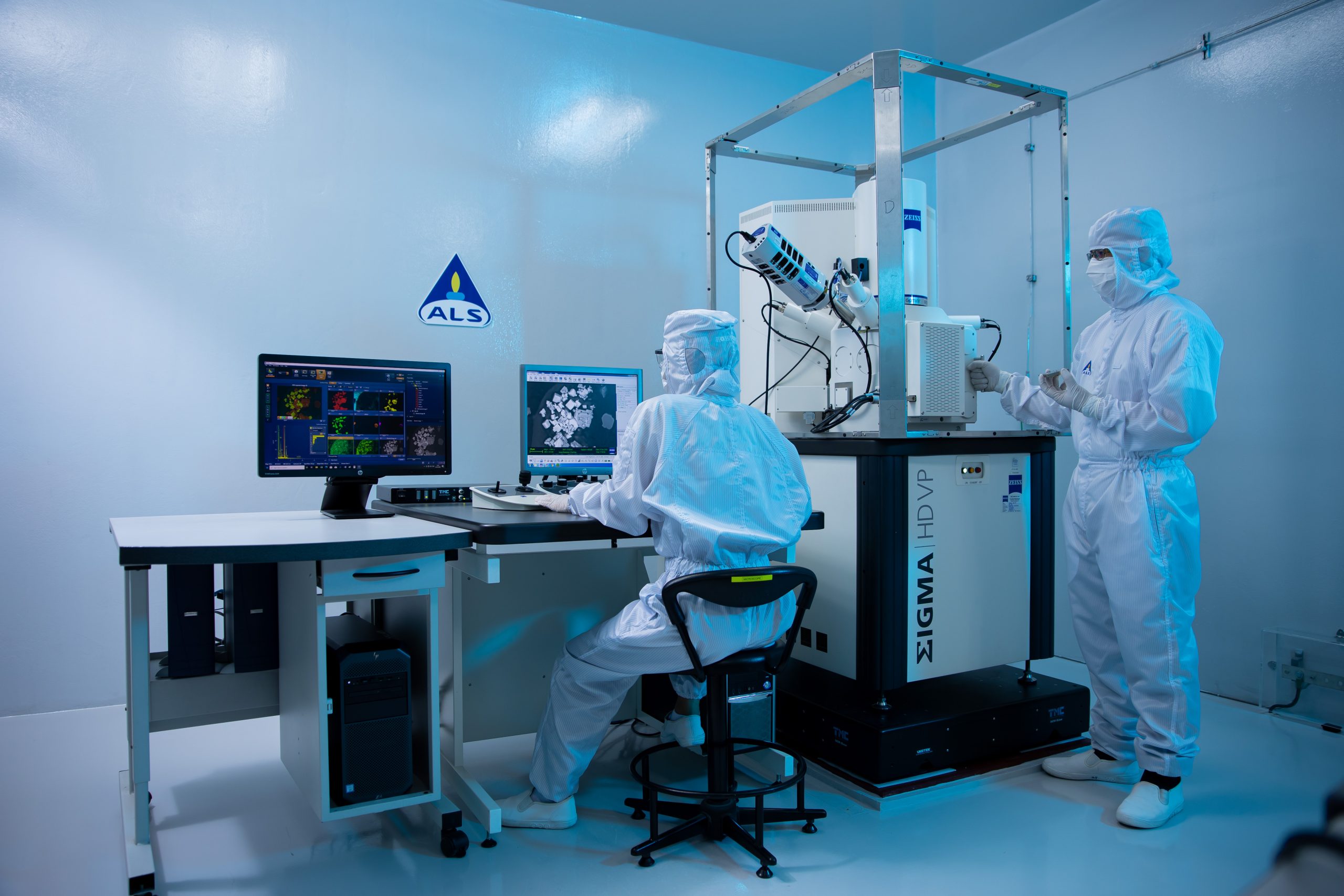 Two people wearing cleanroom suits operate a Sigma HD VP microscope and computer workstations inside a sterile laboratory with ALS branding.