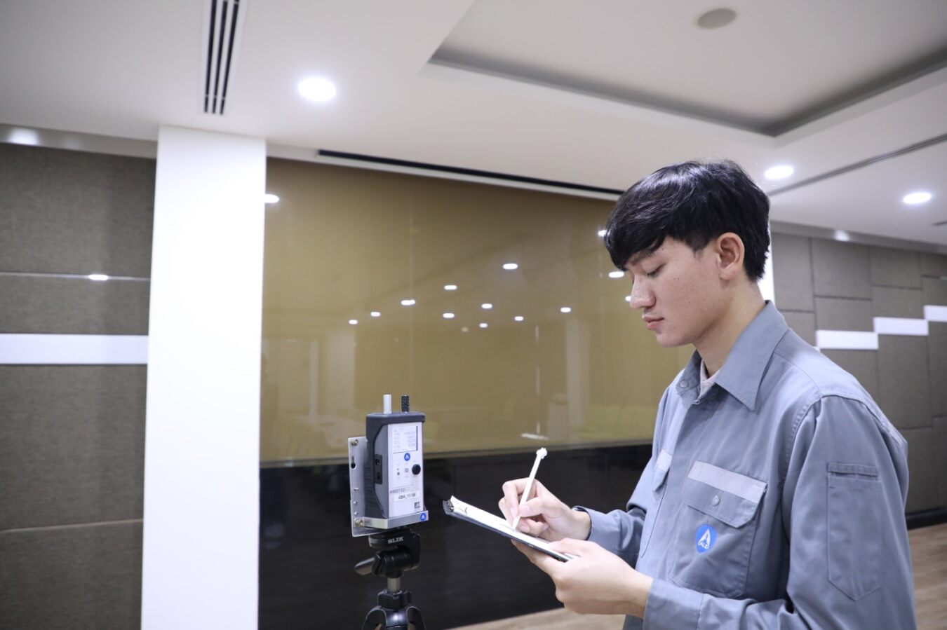 A person in a work uniform records measurements on a tablet or clipboard next to an indoor air quality monitoring device mounted on a tripod inside an indoor workspace.