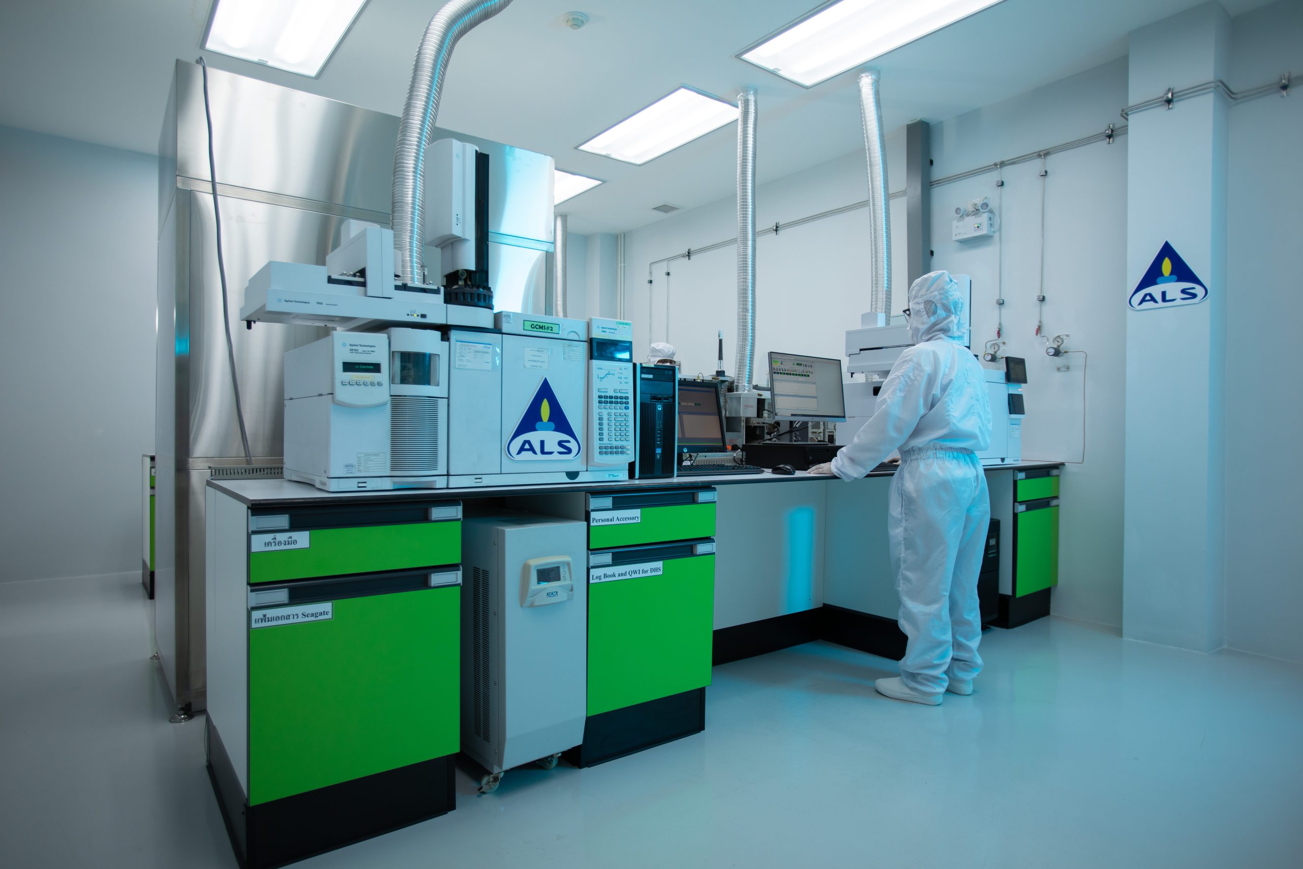 A person wearing a full cleanroom suit operates advanced laboratory analysis equipment in a bright, sterile testing laboratory, with green cabinets and ALS-branded instruments.