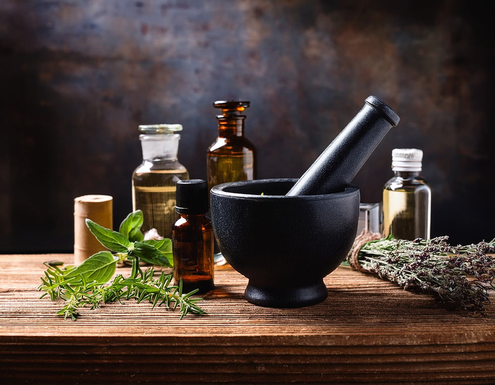 Herbal medicine setup on a wooden table, featuring a black mortar and pestle surrounded by fresh herbs, dried plants, and various small glass bottles filled with oils or extracts.