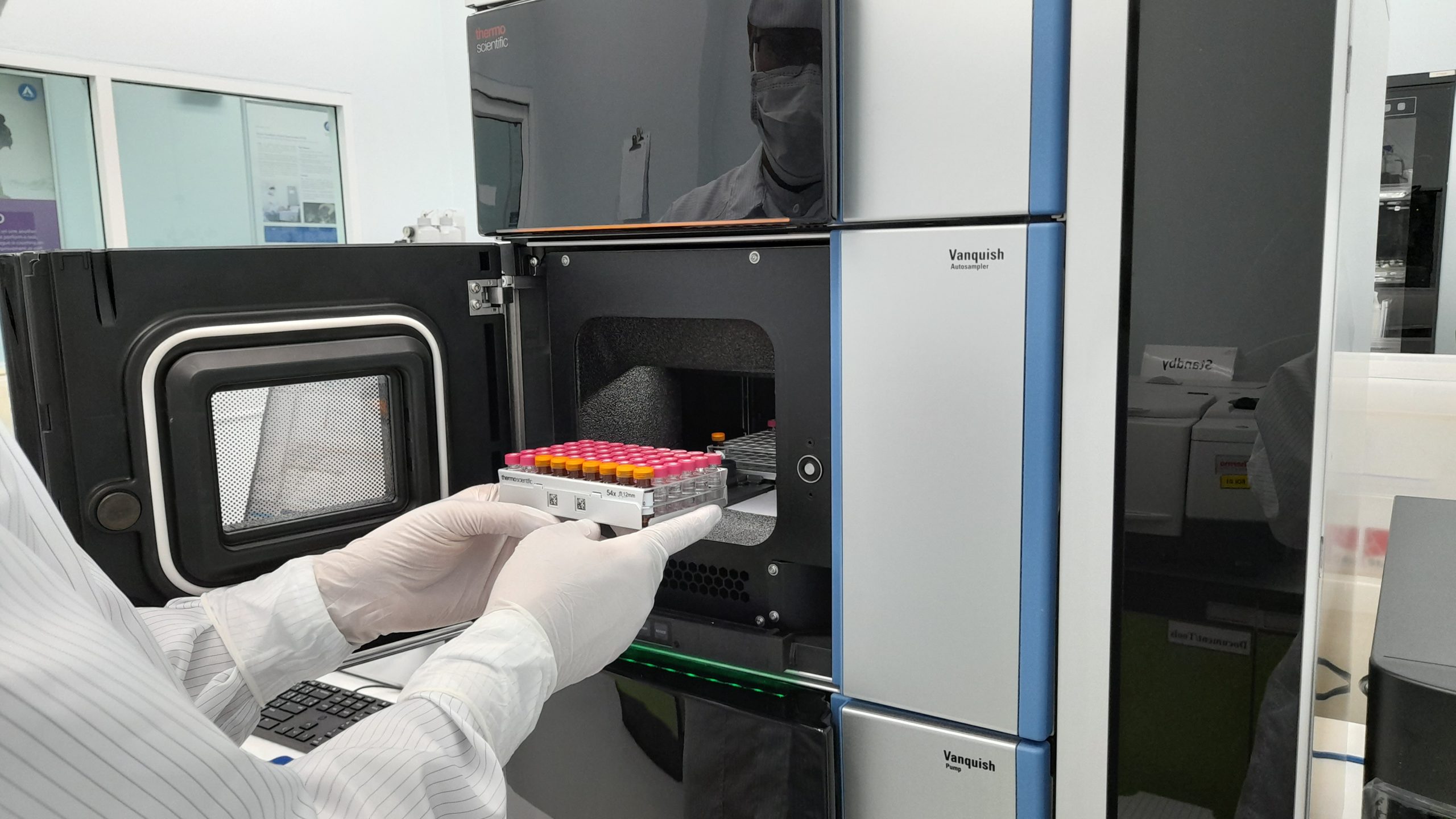 Gloved hands place a rack of sample vials into a high‑performance liquid chromatography (HPLC) system inside a clean laboratory environment.