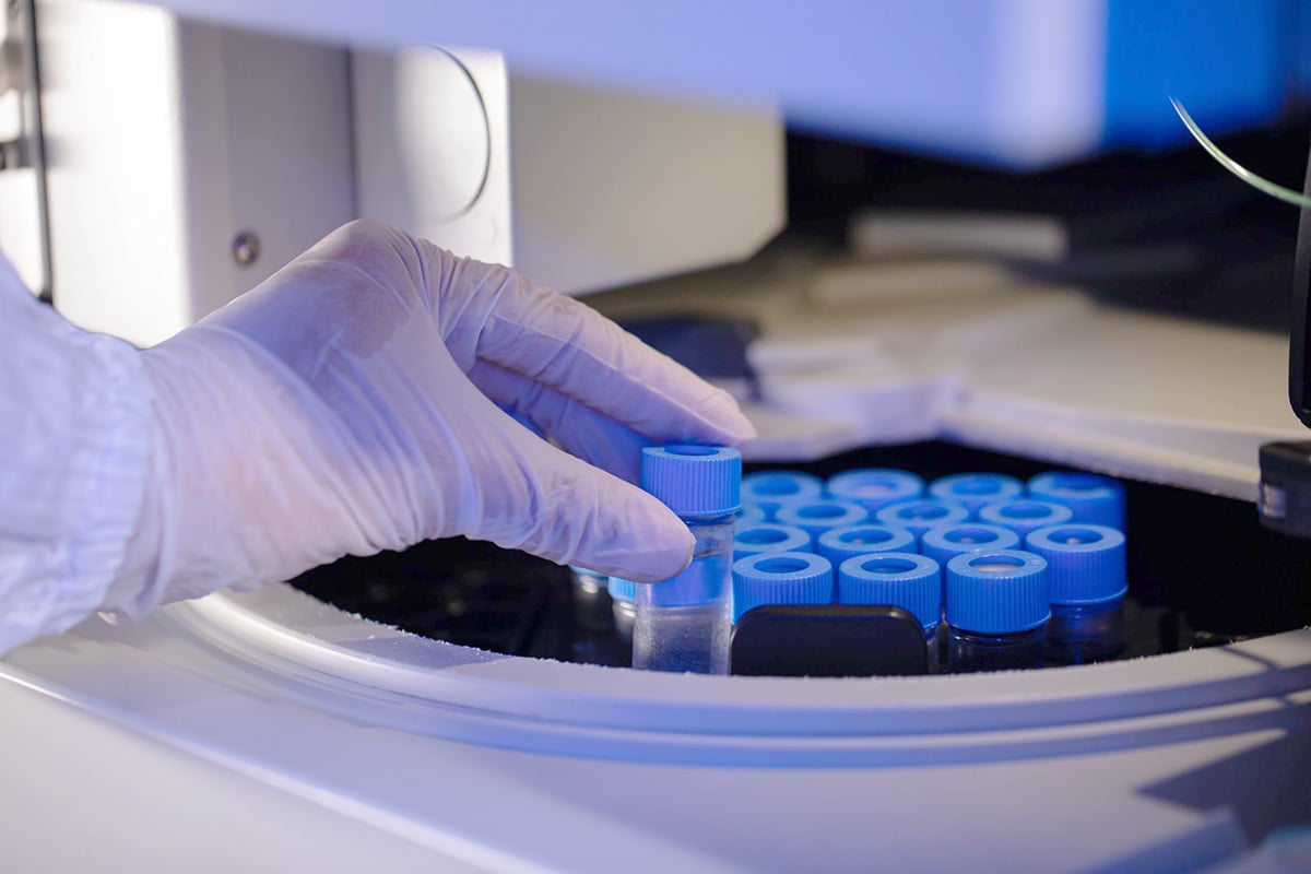 A gloved hand places a blue‑capped sample vial into an autosampler tray inside an ion chromatography laboratory instrument.