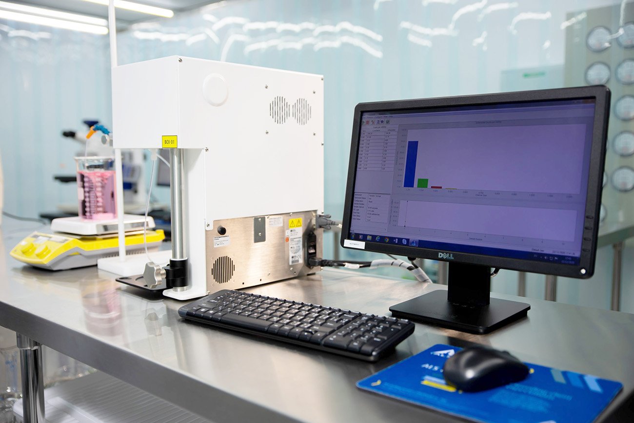A liquid‑borne particle counting instrument connected to a computer displays analysis results on a laboratory workbench, with a beaker of pink liquid visible in the background.