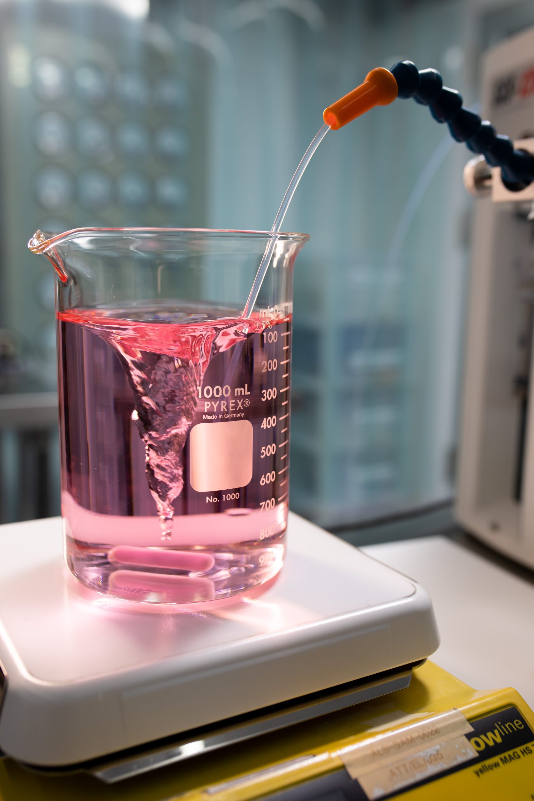 A laboratory beaker containing pink liquid sits on a hot plate while a tube dispenses liquid into the beaker for chemical testing or analysis.