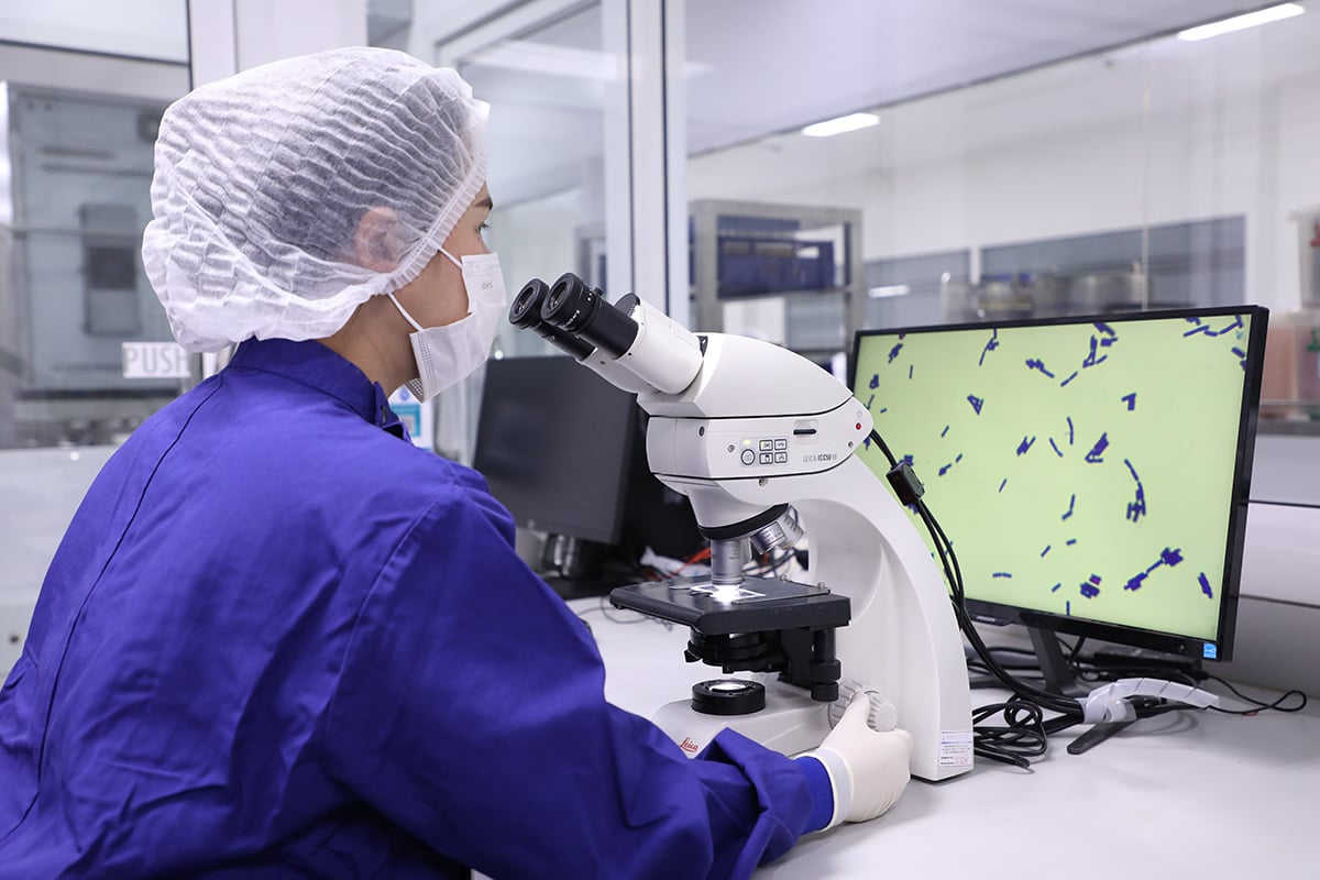 A person wearing protective laboratory clothing uses a microscope at a workstation, with a computer monitor displaying magnified microorganisms in a controlled laboratory environment.