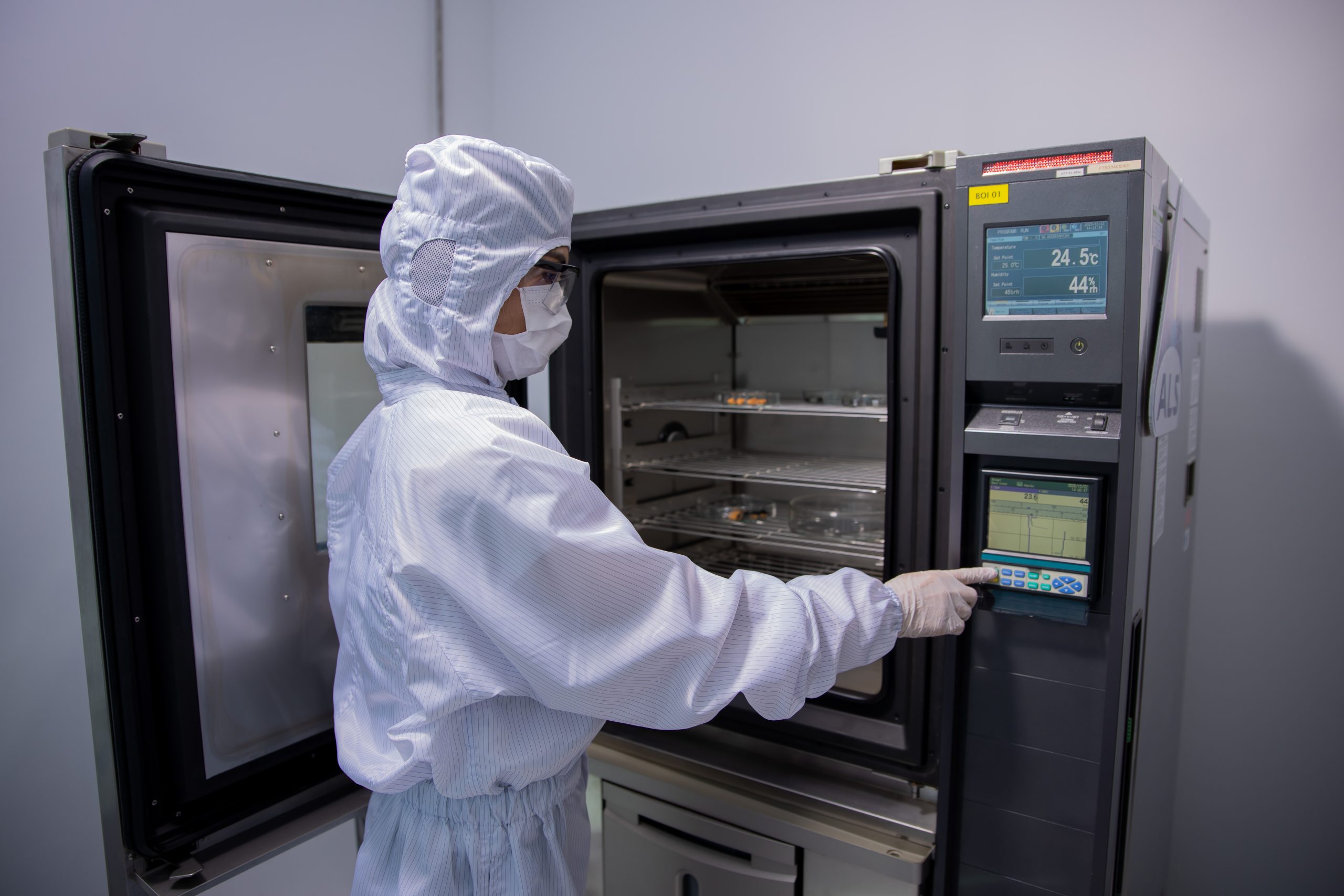 A person wearing a cleanroom suit adjusts settings on a temperature and humidity test chamber with an open door, using a digital control panel in a laboratory environment.