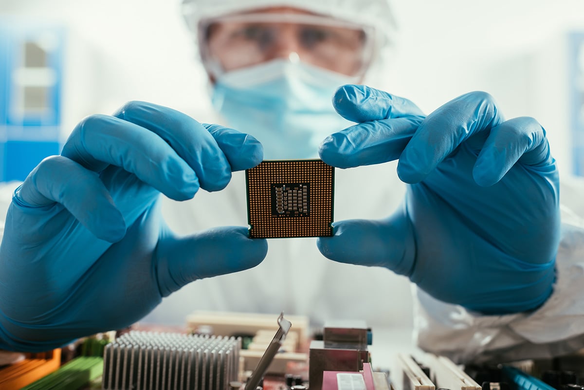 Close-up of a person wearing blue nitrile gloves and protective lab clothing, holding a small square computer processor chip between both hands. The processor is positioned above an exposed computer motherboard with visible components and circuitry. The background is softly blurred, suggesting a clean laboratory or electronics testing environment.