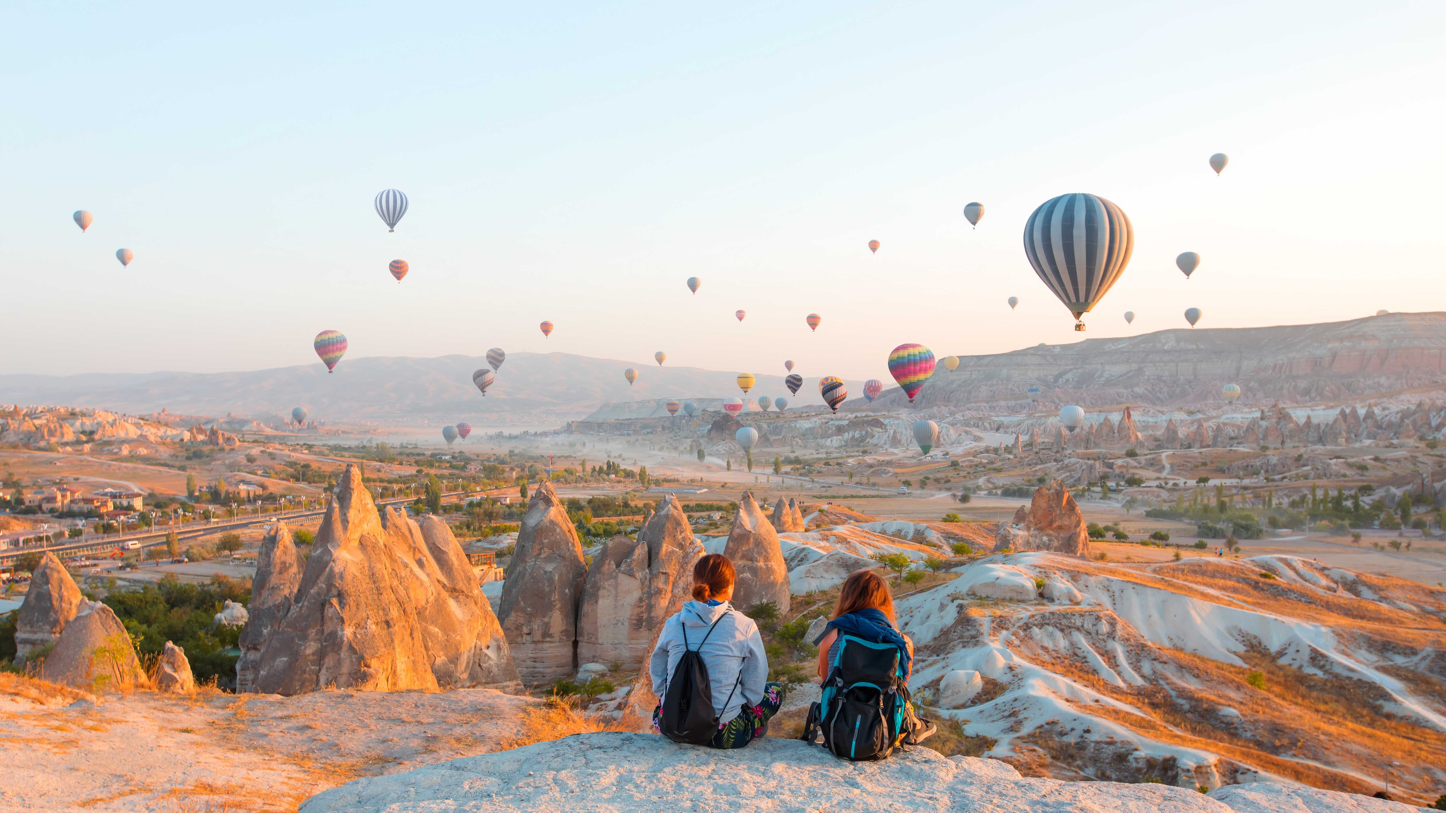 Moleküler Kirlilik ve Doğal Manzaranın Kesişimi Two people sitting on a rocky ledge overlooking Cappadocia’s unique rock formations at sunrise, with numerous colorful hot air balloons floating in the sky.