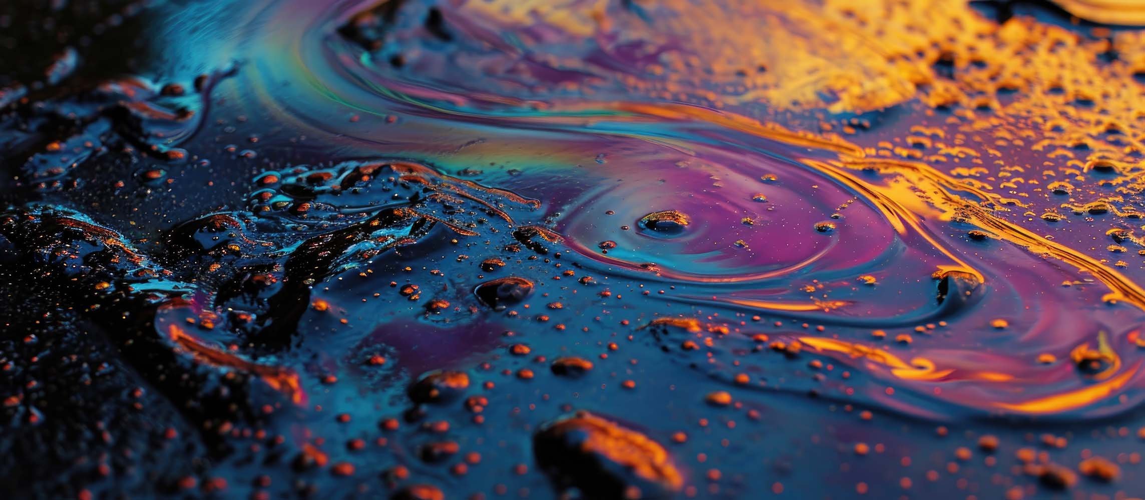 Close-up of an oil spill on a dark surface, showing swirling patterns with vibrant iridescent colors including blue, purple, orange, and yellow.