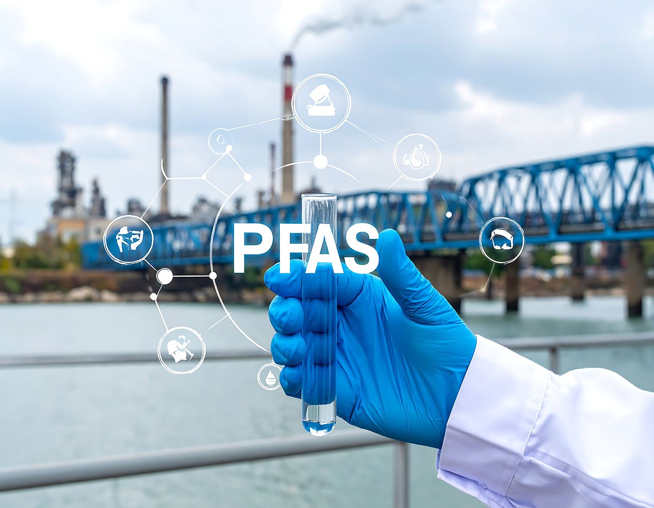 A gloved hand holds a test tube with clear liquid in front of an industrial site and bridge, with the text “PFAS” and chemical icons overlaid.