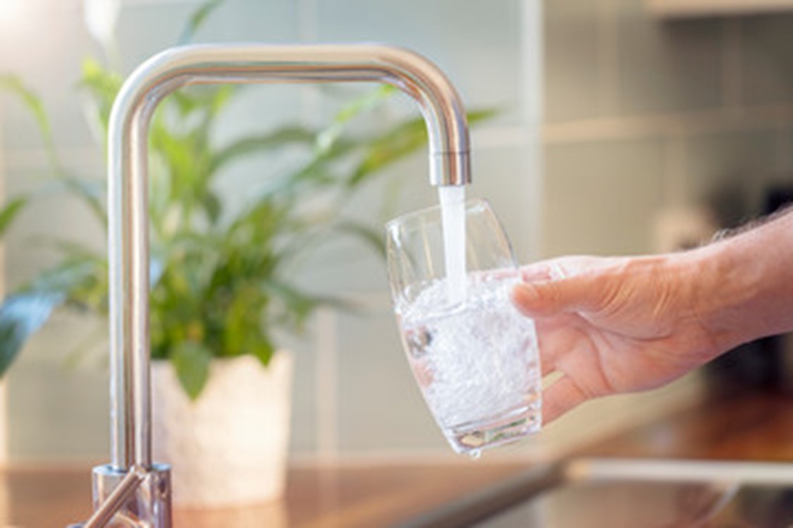 Filling a Glass with Water A hand holding a glass under a running kitchen faucet, filling the glass with water. There is a plant in the background on the left side of the image.