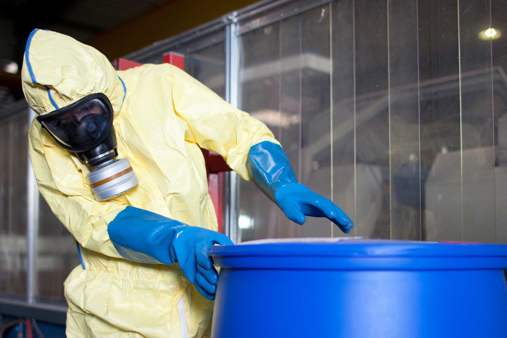 A person wearing a hazmat suit and gloves approaches a blue barrel for testing.