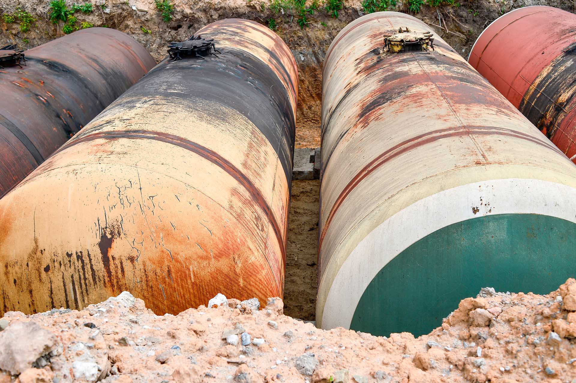 Three large rusty pipes are laid in a trench.