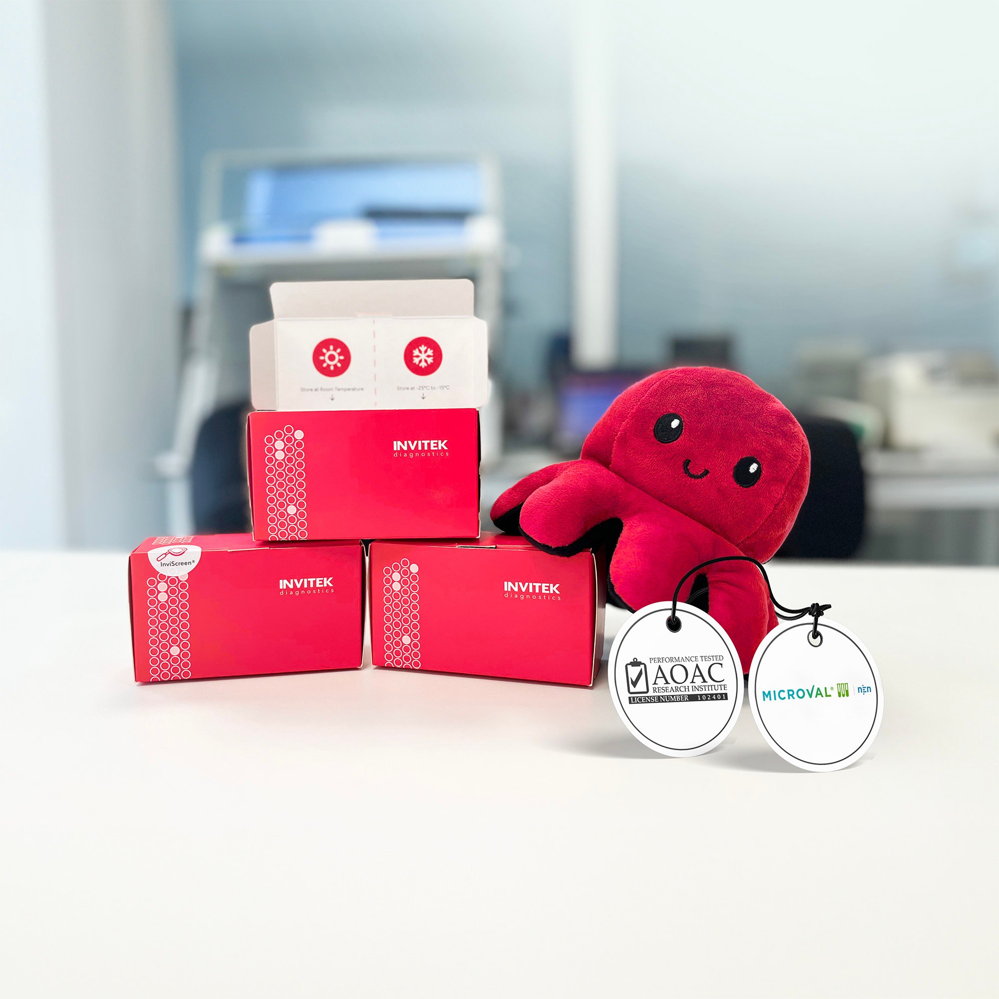 Three red and white boxes labeled 'INVITEK' stacked on a table, with an open box on top revealing its contents, next to a plush toy resembling an octopus with two medals.