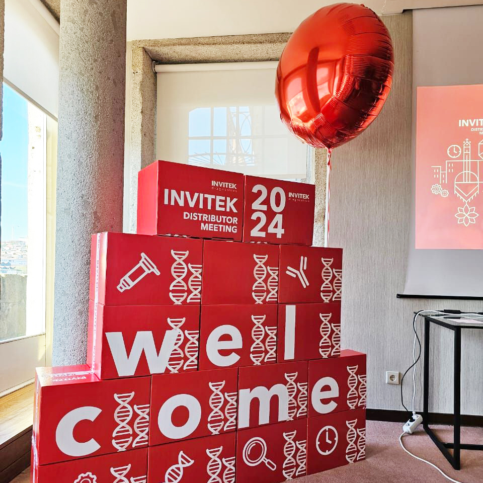Stack of red boxes spelling out 'welcome' with a large red balloon on top, displaying 'INVITEK DISTRIBUTOR MEETING 2024' and scientific illustrations.