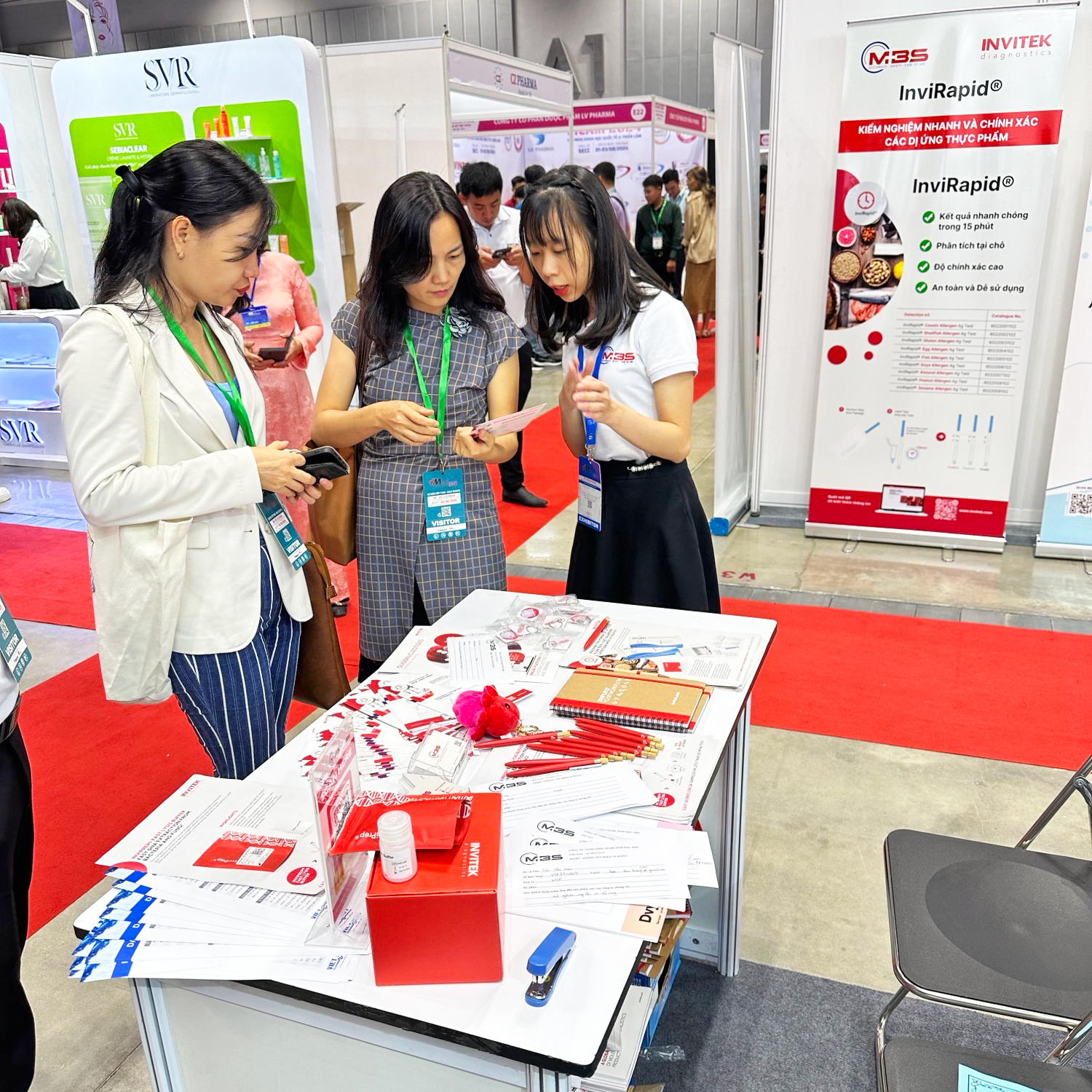Booth at a convention with four people around a table with promotional materials, banners, and posters in Vietnamese advertising medical products.