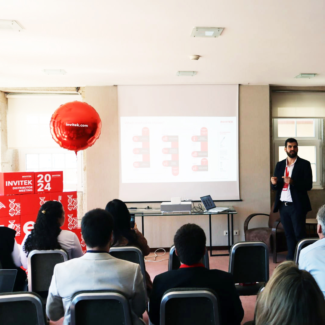 Conference room with attendees facing a presenter near a projection screen, with a red balloon and banner reading 'INVITEK 2024.