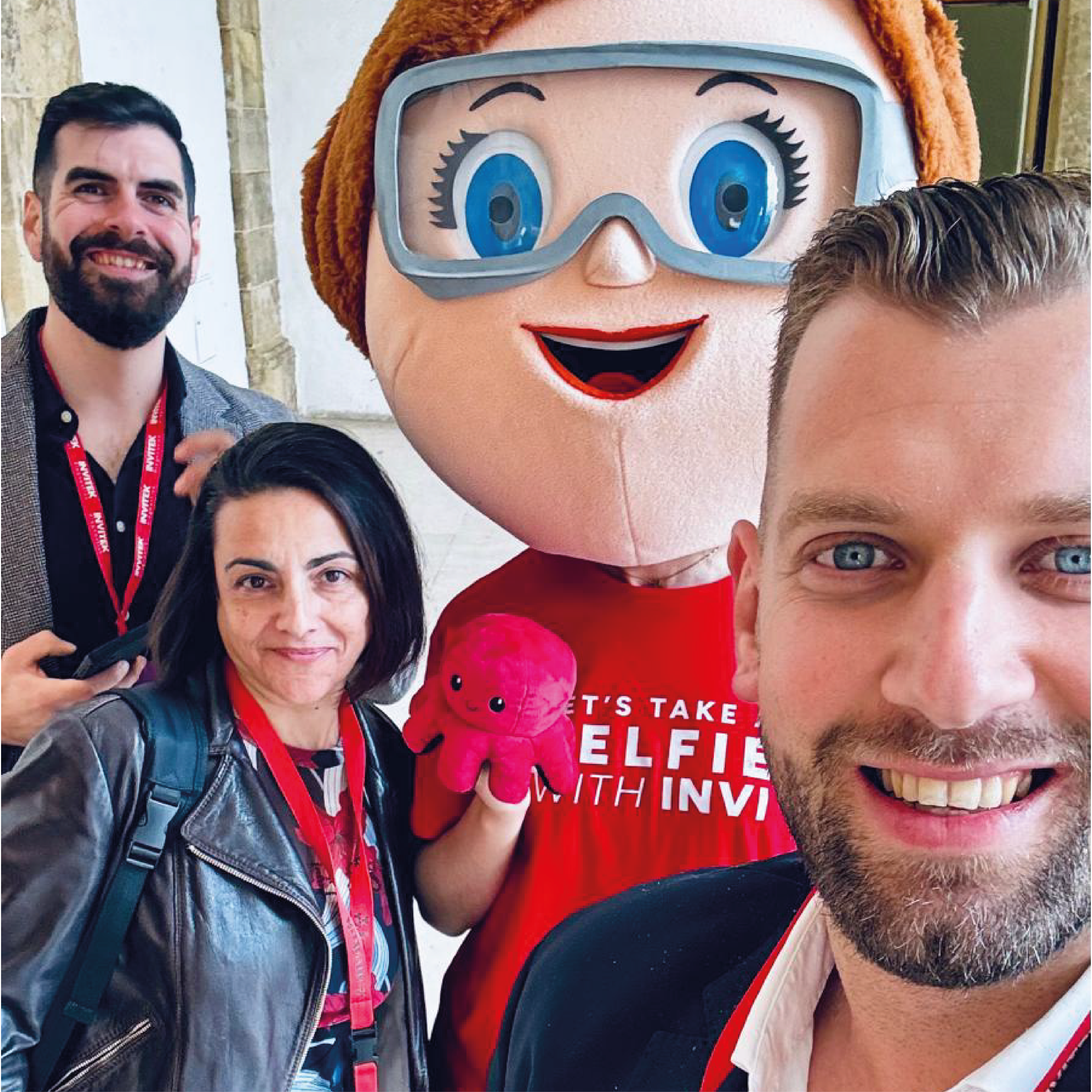 Group of people posing for a photo, with one person holding a small pink plush toy and another wearing a red shirt that reads 'LET'S TAKE A SELFIE WITH INVI.' All individuals have red lanyards with badges.