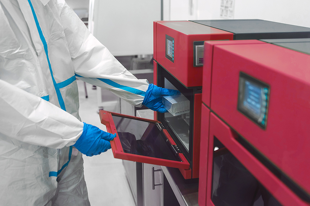 Technician in full PPE and blue gloves loading a tray into a red PCR analyzer